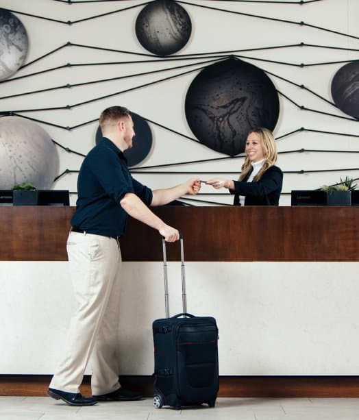 Man with white pants and black shirt at hotel reception checkin counter, holding a carry-on suitcase